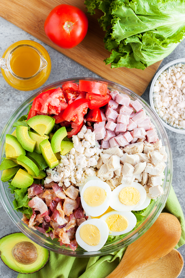 Birds-eye view of cobb salad in a bowl surrounded by serving utensils, avocado, tomatoes, lettuce, and cheese. 