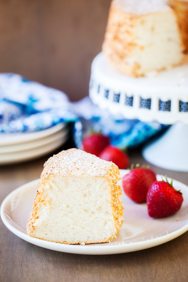 Slice of angel food cake on a plate with fresh strawberries and angel food cake on a cake stand in the background.  