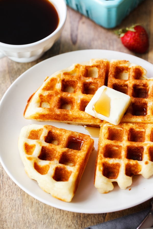Yeast waffle on a plate with a pat of butter surrounded by a mug of coffee and fresh strawberries. 