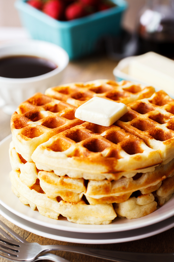 Stack of yeast waffles on a plate topped with pat of butter and coffee, fresh strawberries, butter, and syrup in the background. 