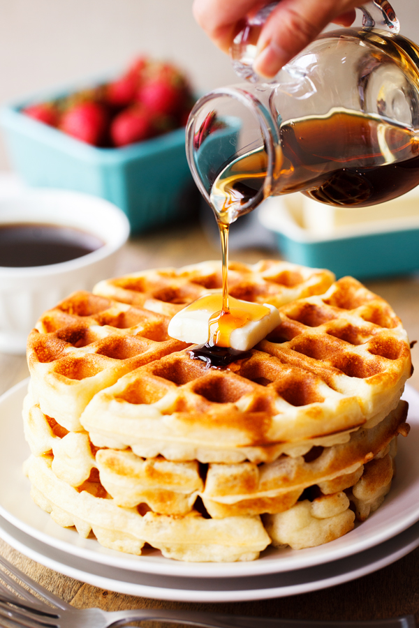 Maple syrup being poured over a stack of yeast waffles with fresh strawberries, coffee, and butter in the background. 