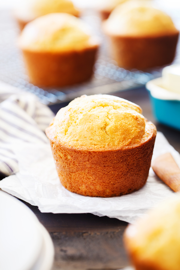Cornbread muffin on layers of parchment paper with cornbread muffins on a cooling rack and butter in the background. 