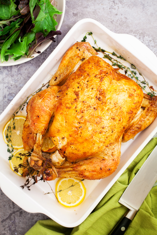 Birds-eye view of roast chicken, sprigs of thyme, and lemon slices in a baking dish and a side salad.