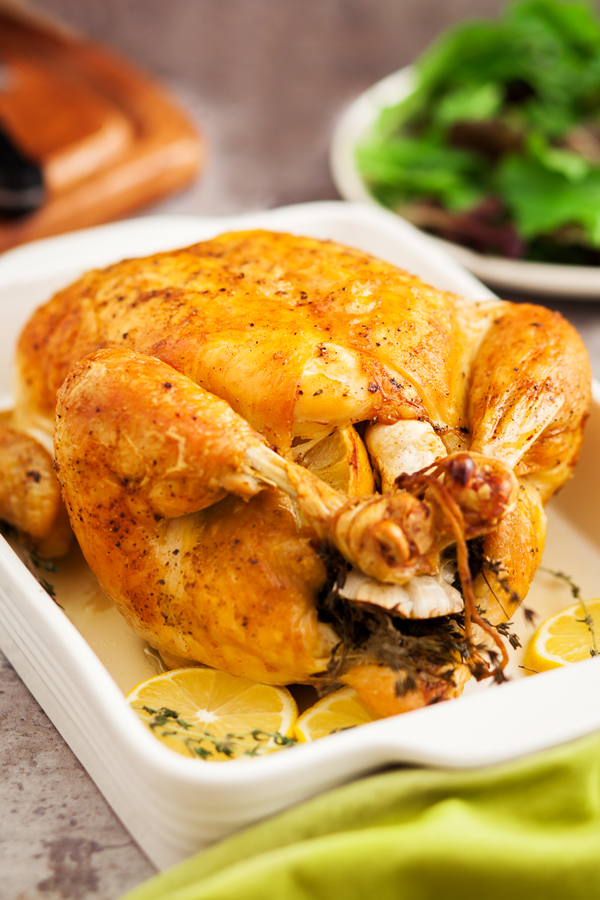 Roast chicken, sprigs of thyme, and lemon slices in a baking dish and a side salad in the background.