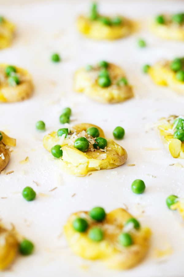 Smashed fingerling potatoes on parchment paper garnished with peas, rosemary, and parmesan cheese. 