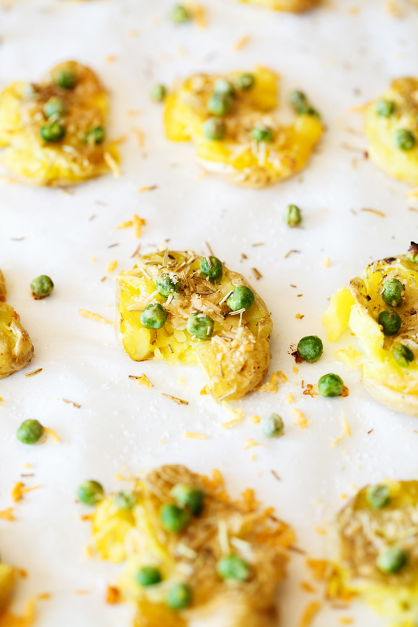 Smashed fingerling potatoes on parchment paper garnished with peas, rosemary, and parmesan cheese. 