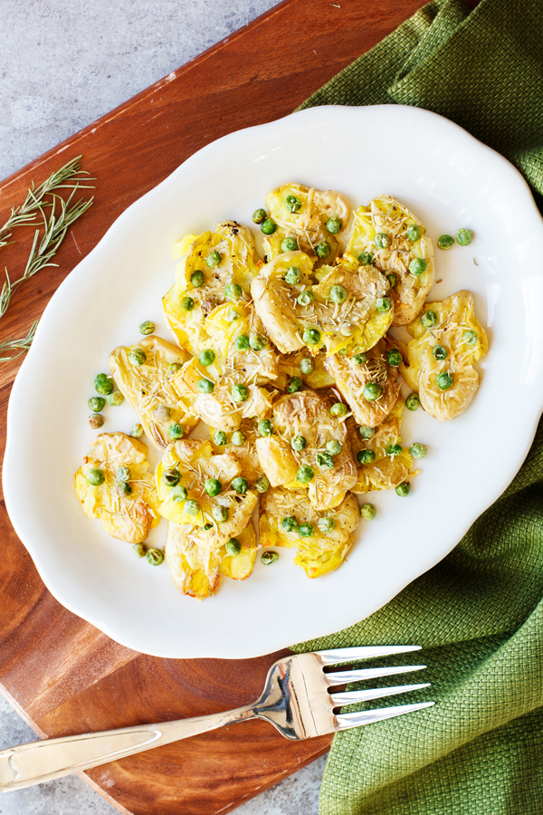 Birds-eye view of smashed fingerling potatoes on a plate garnished with peas, rosemary, and parmesan cheese. 