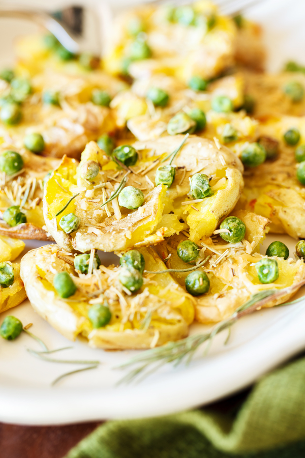 Smashed fingerling potatoes on a plate garnished with peas, rosemary, and parmesan cheese. 