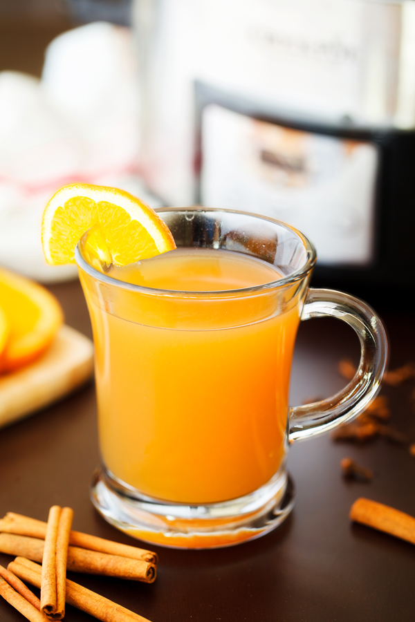 Glass of mulled cider garnished with an orange slice with cinnamon sticks in the foreground and slow cooker and sliced orange garnishes in the background. 