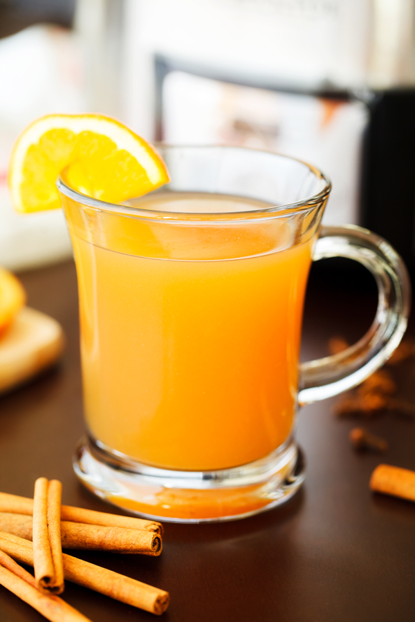 Glass of mulled cider garnished with an orange slice with cinnamon sticks in the foreground and slow cooker in the background. 