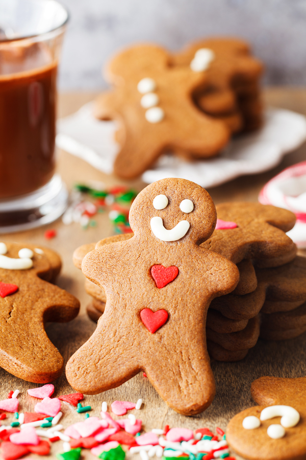 Decorated gingerbread cookie with sprinkles in the foreground and gingerbread cookies and a glass of hot chocolate in the background. 