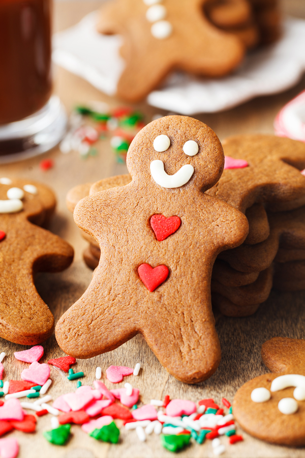 Decorated gingerbread cookie with sprinkles in the foreground and gingerbread cookies and a glass of hot chocolate in the background. 