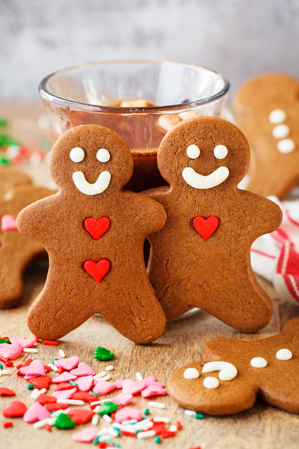 Two decorated gingerbread cookies leaning against a glass of hot chocolate with sprinkles in the foreground and gingerbread cookies in the background. 