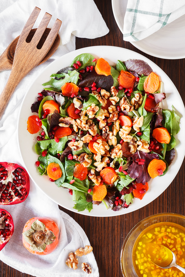 Birds-eye view of a persimmon and pomegranate salad surrounded by fresh pomegranate, persimmon, dressing, and serving utensils.