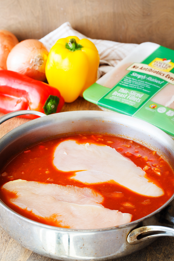 Raw chicken in a pan of red sauce with bell peppers, onions, and Foster Farms chicken in the background. 