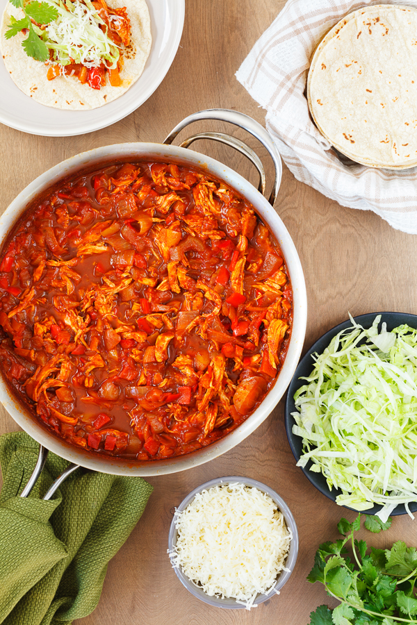 Birds-eye view of shredded chicken in a red sauce in a pan surrounded by an open-face taco, tortillas, shredded lettuce, cilantro, and shredded cheese.