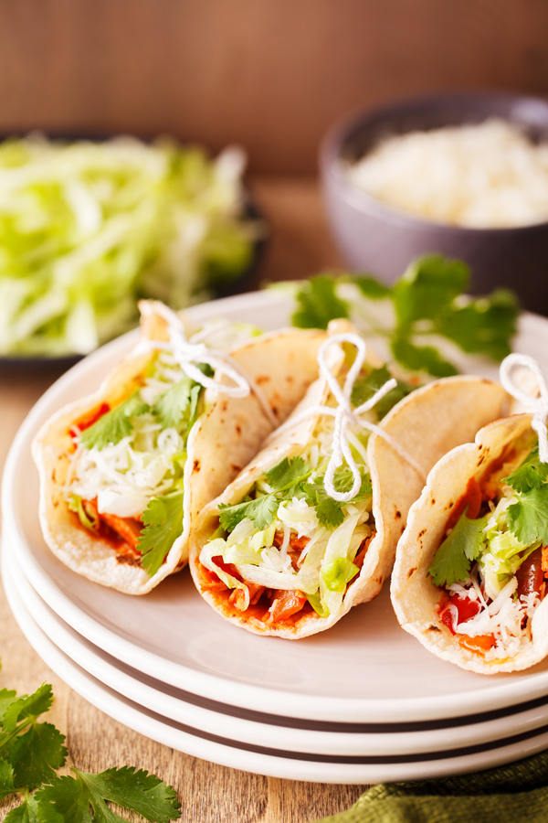 Three chicken ropa vieja tacos tied with string on a plate with lettuce and shredded cheese garnishes in the background. 