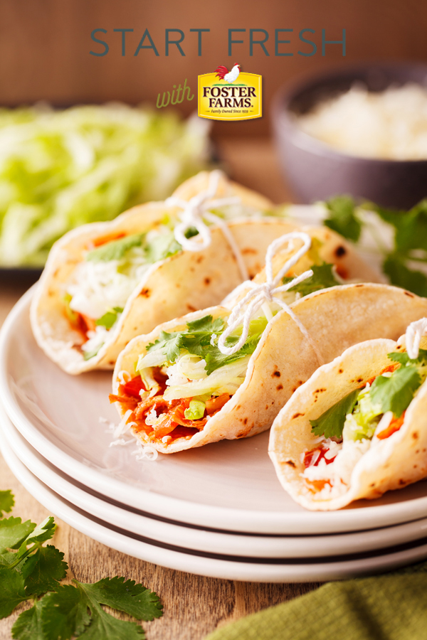 Three chicken ropa vieja tacos tied with string on a plate with lettuce and shredded cheese garnishes in the background. 