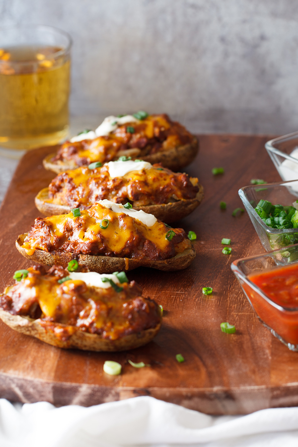 Four Chili Cheese Stuffed Potato Skins with salsa, scallions, and sour cream garnishes on the side and beer in the background. 