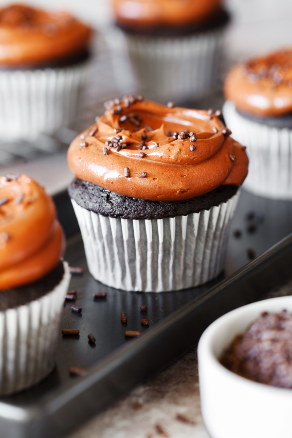 Eye-level view of three chocolate cupcakes frosted with chocolate frosting with chocolate sprinkles on a serving dish and chocolate cupcakes on a cooling rack in the background. 