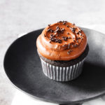 Chocolate cupcake frosted with chocolate frosting and chocolate sprinkles on a plate.