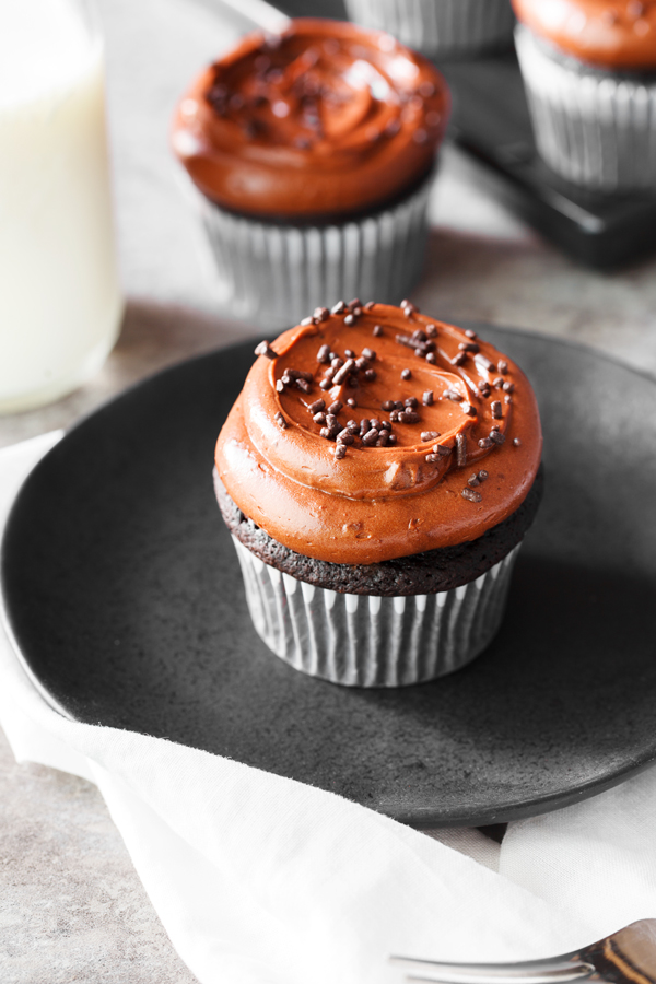 Chocolate cupcake frosted with chocolate frosting and chocolate sprinkles on a plate and cupcakes and milk in the background. 