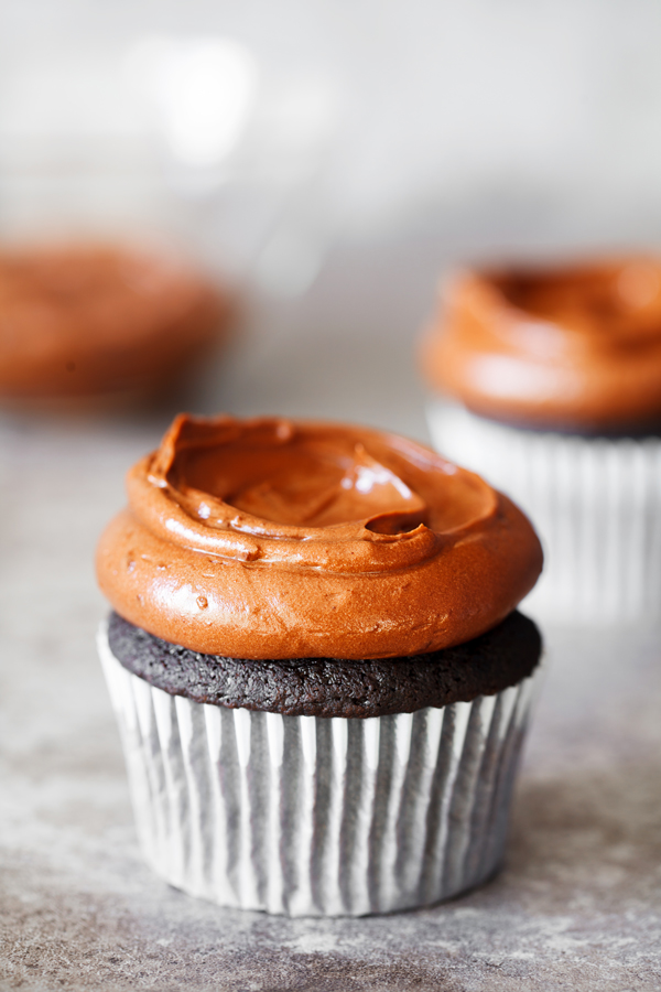 Eye-level view of chocolate cupcake frosted with chocolate frosting with frosted chocolate cupcake and bowl of frosting in the background.