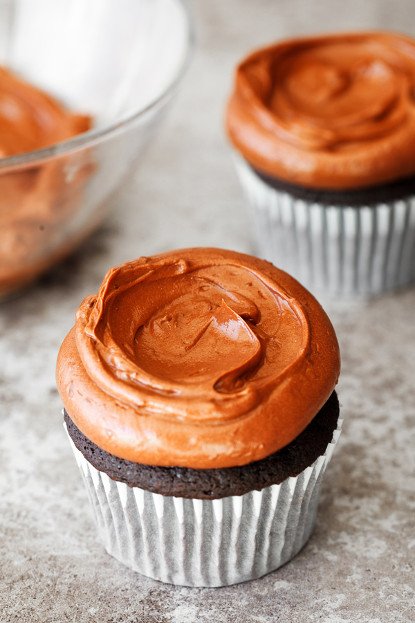 Chocolate cupcake frosted with chocolate frosting with frosted chocolate cupcake and bowl of frosting in the background.