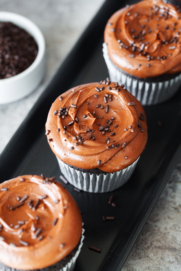 Three chocolate cupcakes frosted with chocolate frosting on a serving dish and sprinkled with chocolate frosting. Small bowl of chocolate sprinkles in the background.