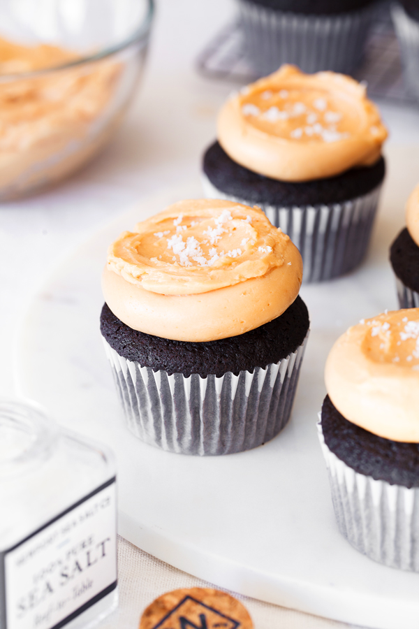 Chocolate cupcakes with dulce de leche frosting and a sprinkling of sea salt on a serving platter. Bowl of dulce de leche frosting in the background and sea salt in the foreground. 