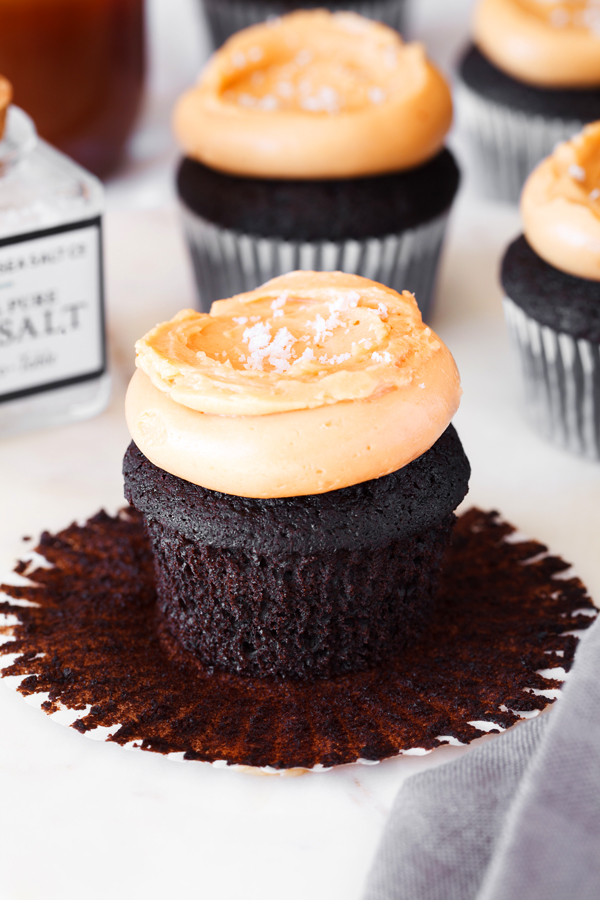 Chocolate cupcake with dulce de leche frosting and a sprinkling of sea salt and paper liner peeled off. Sea salt and cupcakes in the background. 