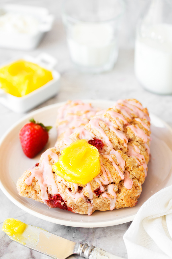 Strawberry scone with a dollop of lemon curd on a plate and lemon curd, cream cheese, and milk in the background. 
