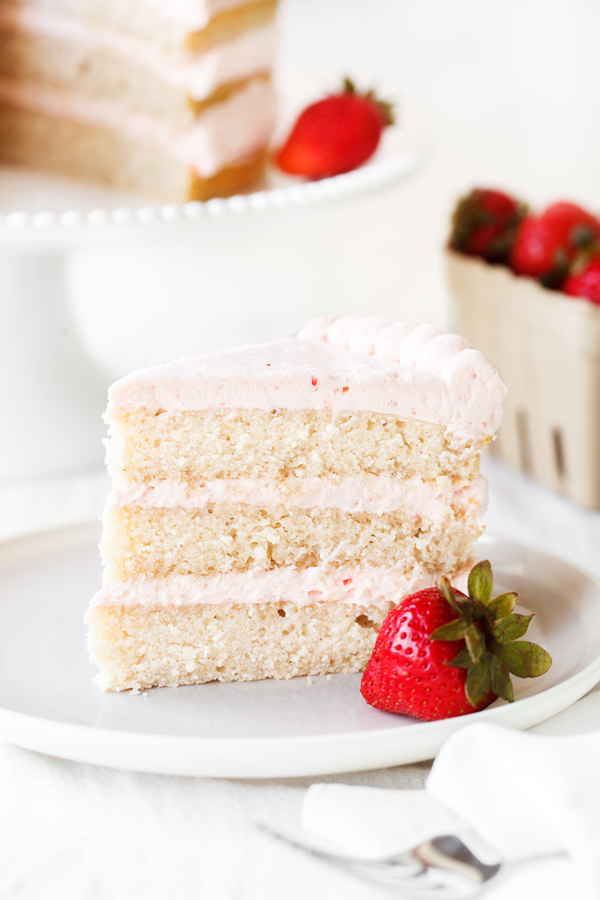 Slice of 3-layered strawberry cake in the foreground and a whole strawberry cake on a cake stand in the background. 
