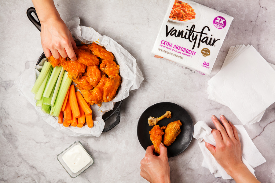 Hands reaching for buffalo wings on counter top and using Vanity Fair® Extra Absorbent Napkins.